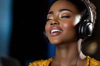 Young African American woman wearing headphones and yellow sweater smiling with eyes closed, enjoying music against dark blue background with bokeh lights.