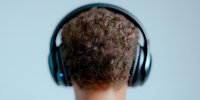 Back view of African American male with curly hair wearing wireless blue headphones against light background, focus on natural hair texture and modern audio device.