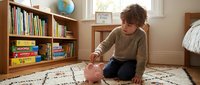 A young child with curly brown hair, appearing to be of mixed ethnicity, sits on a patterned rug in a bright, organized bedroom. The child wears a beige sweater and dark pants while playing with a pink piggy bank, learning about saving money and financial literacy at home.