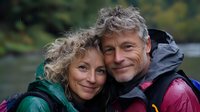 Mature Caucasian couple smiling outdoors in hiking gear, sharing intimate moment. Woman with curly blonde hair and man with gray hair wearing waterproof jackets embrace in nature.