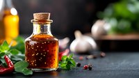 Small glass bottle with spicy chili oil, fresh herbs, garlic and red peppers on dark rustic background. Food photography for cooking blogs and recipe websites.
