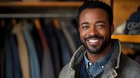 Young African American man with bright smile wearing casual denim shirt and cardigan standing in clothing store, genuine cheerful expression for retail and fashion marketing.
