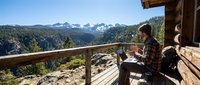 Young man in plaid shirt relaxing on wooden mountain cabin deck with panoramic alpine vista, enjoying peaceful morning coffee break in nature retreat setting.