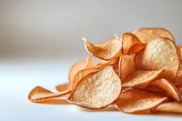 Crispy golden potato chips scattered on white surface with soft focus and shallow depth of field, creating appetizing snack food composition.