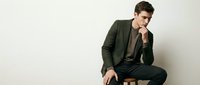 Young handsome Caucasian businessman in elegant dark green suit sitting thoughtfully with hand on chin, professional corporate portrait on neutral background.