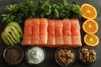Fresh raw salmon fillet portions with avocado, oranges, kale, walnuts and seasonings arranged on dark stone background for healthy cooking preparation.