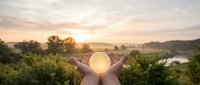 Hands holding glowing sun orb at sunset over rural landscape with river and trees, symbolizing hope, success, and renewable energy for environmental and business concepts.