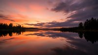 Serene lake at sunset with dramatic orange and purple sky reflecting on calm water, surrounded by silhouetted evergreen trees along shoreline.