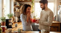 Young mixed-race woman laughing while bearded Caucasian man offers her red mug in cozy kitchen. Couple enjoying morning coffee together at home.