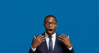 Shocked African American male professional wearing glasses and dark business suit with striped tie gestures with open hands against solid blue background, expressing surprise or excitement.