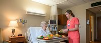 Friendly female nurse in pink scrubs serving healthy meal to elderly patient in modern hospital room with warm lighting and comfortable amenities.