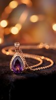 Silver pendant necklace with purple teardrop gemstone on dark surface, warm bokeh lights background. Luxury jewelry for fashion, accessories, and elegant styling.