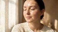 Middle-aged woman with dark hair and fair complexion wearing beige knit sweater, eyes closed in peaceful expression, natural window lighting creating soft shadows on face, serene indoor portrait.