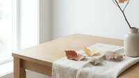 Autumn leaves scattered on white linen fabric over wooden table near window with natural light creating cozy seasonal home decor atmosphere.