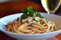 Fresh spaghetti pasta garnished with parsley and black pepper served in white ceramic bowl, with glass of white wine in soft background lighting.