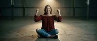 Young Caucasian woman celebrating success sitting cross-legged on wooden floor in empty warehouse with raised fists showing joy and achievement.