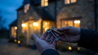 Hands holding house keys in front of blurred home with warm lights at dusk, symbolizing homeownership, real estate success, and new property investment.