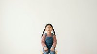 Young Asian girl with braided hair wearing denim overalls and pink long-sleeve shirt, sitting and looking upward with cheerful expression against neutral background.