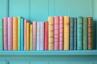 Colorful vintage books arranged on turquoise shelf, creating vibrant rainbow display of pastel and muted tones against matching wall background.