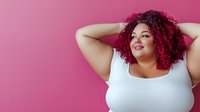 Confident plus size woman with vibrant red curly hair posing against pink background, hands behind head in relaxed, empowering stance.