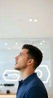 Young man with dark hair and facial hair, appearing to be of Latin or Mediterranean ethnicity, wearing a navy blue button-up shirt, looking upward thoughtfully in a modern office environment with glass panels and ambient lighting.