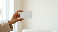 Close-up of male hand holding blank white business card against neutral beige wall with natural window light, suitable for corporate branding and identity mockups.