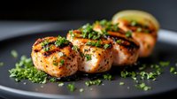 Grilled salmon medallions garnished with fresh herbs and citrus on dark plate, close up view showing juicy texture and caramelized surface.