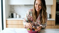 Young Hispanic woman in floral blouse preparing healthy breakfast bowl with fresh berries in modern kitchen, smiling while cooking nutritious meal.