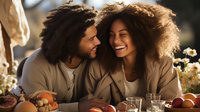 Young mixed race couple sharing intimate moment at dinner table with fresh fruits and flowers, laughing together in warm evening light. Natural lifestyle setting.