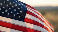 American flag waving outdoors at sunset, stars and stripes close-up with warm golden light and natural landscape background.