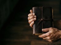 Male hands holding aged leather journal tied with twine against dark background, suitable for publishing, education, storytelling, antique collections, and heritage content.