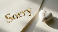 Gold embossed sorry text on cream card beside a cracked white ceramic heart, symbolizing apology, regret, and emotional healing in relationships.
