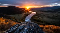 Dramatic sunset over winding river valley with sunburst through mountains, golden autumn colors on hillsides, and rocky overlook creating stunning natural landscape.