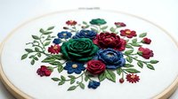 Embroidered floral arrangement featuring red, blue, and green roses with delicate leaves on white fabric stretched in wooden hoop for needlework and craft projects.