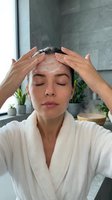 Young woman in white bathrobe applying exfoliating face scrub with closed eyes during skincare routine in modern bathroom spa setting for wellness and beauty.