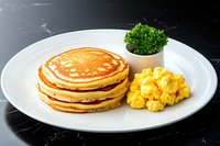 Stack of golden pancakes served on white plate with scrambled eggs and fresh parsley garnish against dark background, classic breakfast presentation.