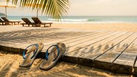 Flip flops on sandy tropical beach with wooden deck, palm tree, ocean waves, and lounge chairs in warm golden sunlight.