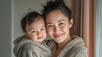 Young Asian mother holding adorable baby in cozy knit sweaters, warm family bonding moment at home with natural window light for parenting content.