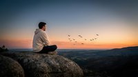 Young man meditating on mountain rock at sunset with birds flying overhead, peaceful mindfulness practice in nature landscape for wellness and spirituality.
