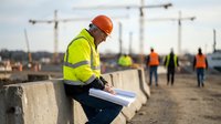 Construction worker in safety gear reviewing blueprints at building site with cranes and team in background for project planning and development.