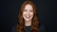 Young Caucasian woman with long wavy red hair smiling warmly at camera wearing gray sweater against dark background for professional headshot portrait.