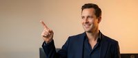 Professional Caucasian businessman in navy suit pointing finger with confident smile against warm gradient background for corporate presentations.