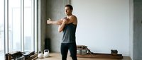 Young athletic Caucasian man stretching arms before workout in modern home gym with kettlebell and large window, active lifestyle concept.