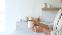Young adult woman with medium skin tone holding white ceramic jar with copper lid in bright kitchen setting, wearing cream cardigan for skincare, cosmetics, wellness product marketing.