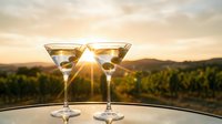 Two elegant martini glasses filled with golden cocktails on outdoor table at sunset with vineyard landscape background creating romantic atmosphere for celebration.
