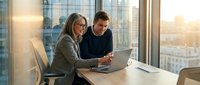 Two professional business colleagues working together on laptop computer in modern office with city skyline view during golden hour sunset light.