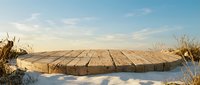 Wooden dock platform extending over frozen winter lake with snow and dry reeds under blue sky. Natural outdoor scene for product placement and display.