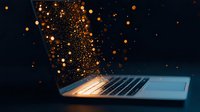 Magical glowing orange particles rising from illuminated laptop keyboard against dark background, creating ethereal technology concept for digital transformation.