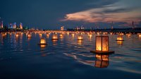Glowing lanterns floating on calm water at twilight with illuminated city skyline in background, creating serene atmosphere for festival celebration.