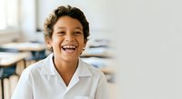 Young boy with dark curly hair and medium skin tone wearing white school uniform shirt, smiling brightly in classroom setting with blurred educational materials background.
